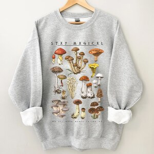 Op de afbeelding: Een grijs sweatshirt met een paddenstoelenillustratie en de tekst "STAY MAGICAL" gedrukt op de voorkant. De illustratie toont een verscheidenheid aan paddenstoelen in verschillende kleuren en vormen.