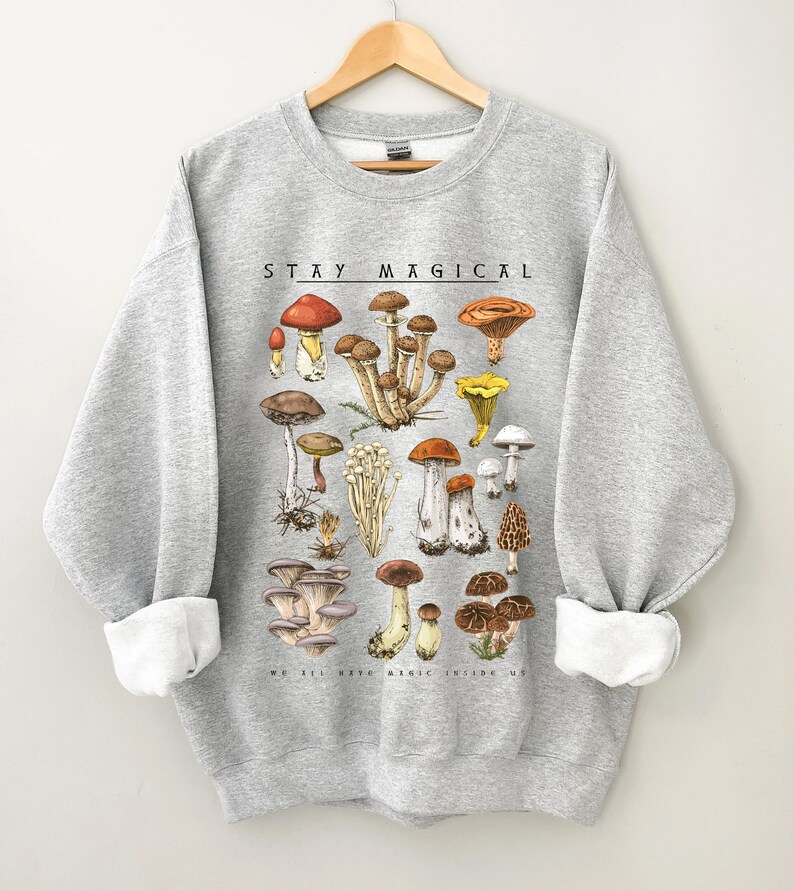 Op de afbeelding: Een grijs sweatshirt met een paddenstoelenillustratie en de tekst "STAY MAGICAL" gedrukt op de voorkant. De illustratie toont een verscheidenheid aan paddenstoelen in verschillende kleuren en vormen.