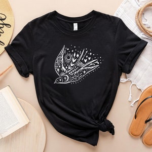May include: Black t-shirt featuring a white graphic of a stylised bird with floral and celestial elements. The bird is surrounded by stars, hearts, and a sun.