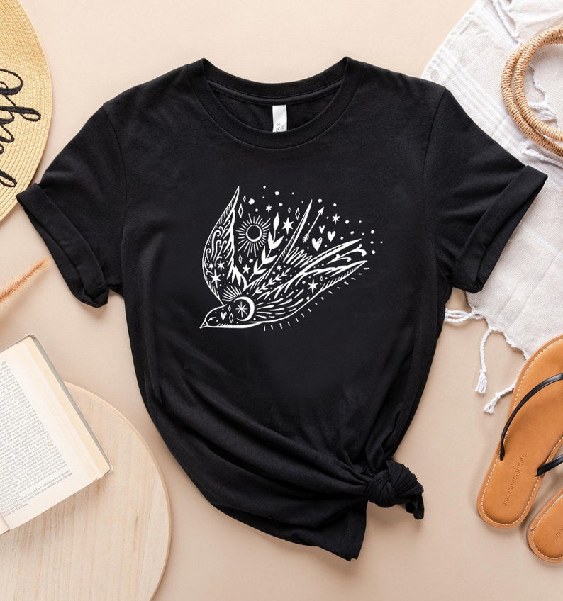 May include: Black t-shirt featuring a white graphic of a stylised bird with floral and celestial elements. The bird is surrounded by stars, hearts, and a sun.