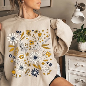 May include: A beige sweatshirt with a floral print featuring yellow, white, and blue flowers. The flowers are arranged in a circular pattern on the front of the sweatshirt.