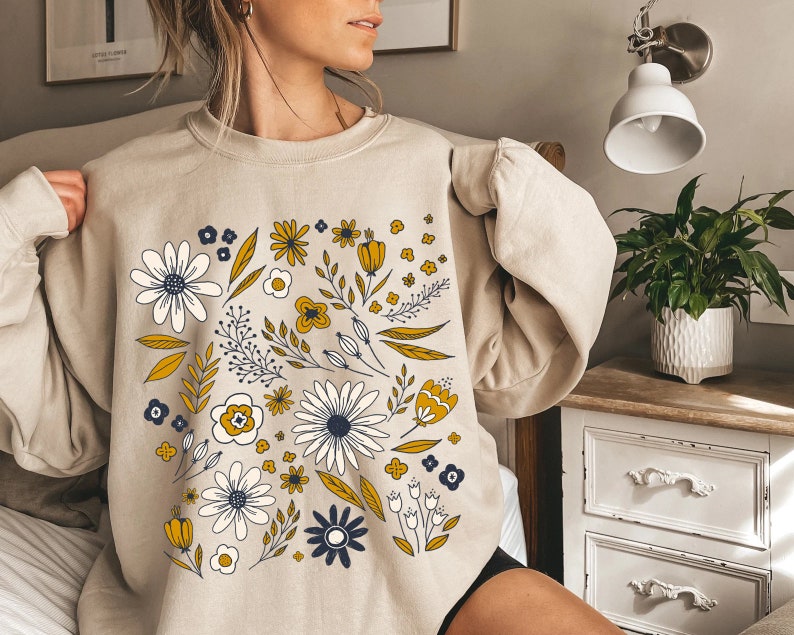 May include: A beige sweatshirt with a floral print featuring yellow, white, and blue flowers. The flowers are arranged in a circular pattern on the front of the sweatshirt.