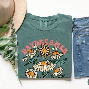 Puede incluir: Una camiseta verde azulada con un gr&aacute;fico retro de flores y el texto "Daydreamer" en rosa.