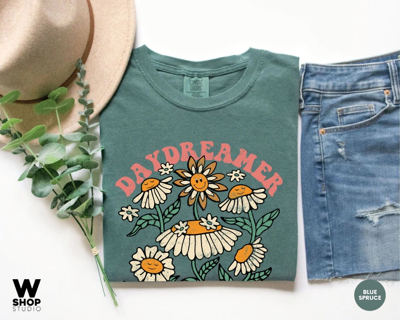 Puede incluir: Una camiseta verde azulada con un gr&aacute;fico retro de flores y el texto "Daydreamer" en rosa.