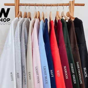 May include: Ten different colored sweatshirts hanging on wooden hangers. The colors are white, ash, sports grey, sand, light pink, light blue, navy, red, maroon, forest green, dark chocolate, and black.