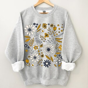 May include: A gray sweatshirt with a floral print in shades of yellow, white, and blue. The flowers are arranged in a circular pattern on the front of the sweatshirt.