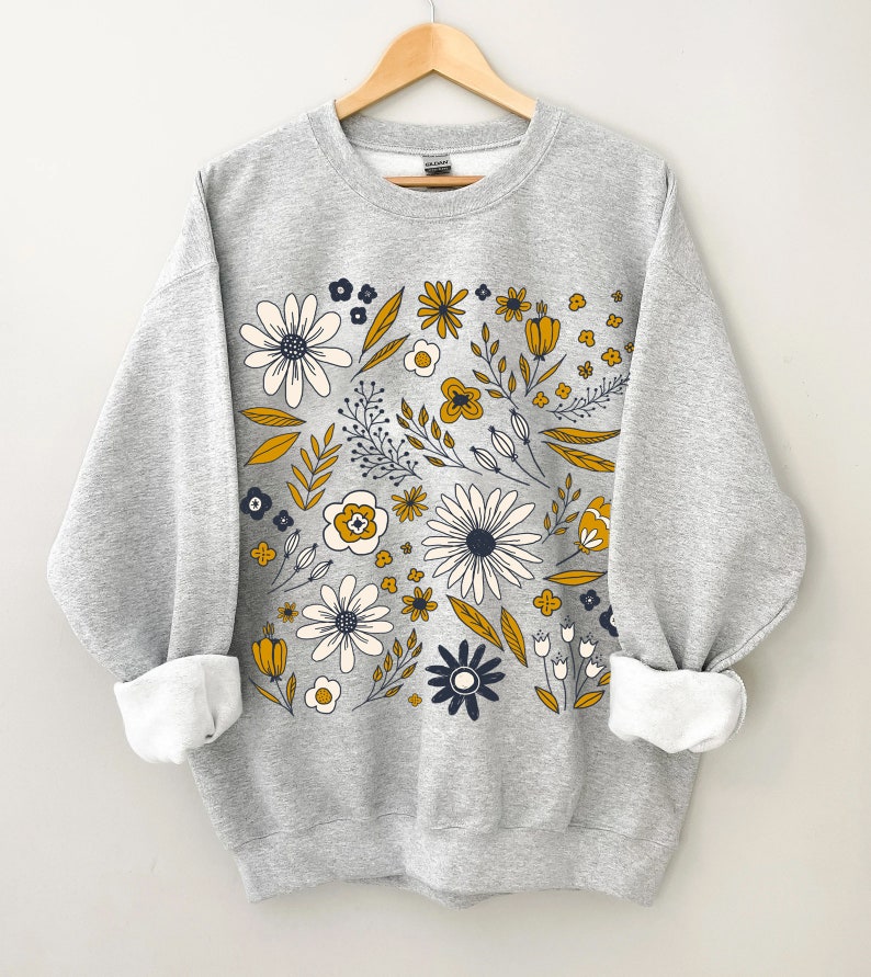 May include: A gray sweatshirt with a floral print in shades of yellow, white, and blue. The flowers are arranged in a circular pattern on the front of the sweatshirt.