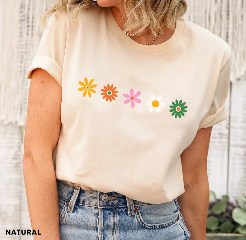 May include: A cream colored t-shirt with a row of five colorful flowers printed on the front. The flowers are yellow, orange, pink, white, and green.
