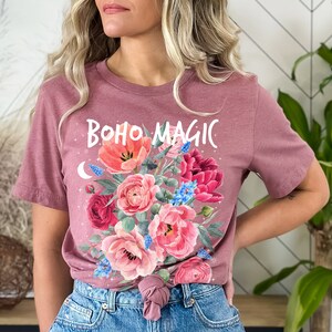 Może przedstawiać: Koszulka w kolorze malwy z kwiatowym wzorem i napisem "boho magic" w białych literach.