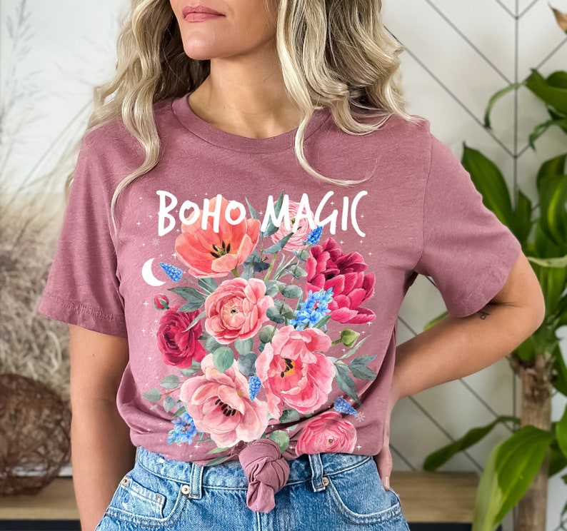 Może przedstawiać: Koszulka w kolorze malwy z kwiatowym wzorem i napisem "boho magic" w białych literach.