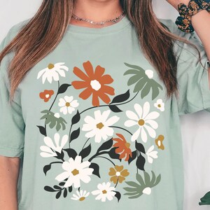 May include: A light green t-shirt featuring a floral design with white, orange, and green flowers. The shirt has a crew neck and short sleeves. The text "W SHOP STUDIO" is at the bottom left.