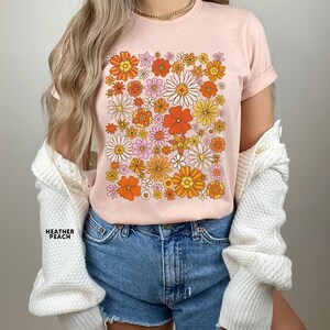 May include: A pink t-shirt with a retro floral print featuring orange, yellow, and pink flowers. The shirt has a crew neck and short sleeves.