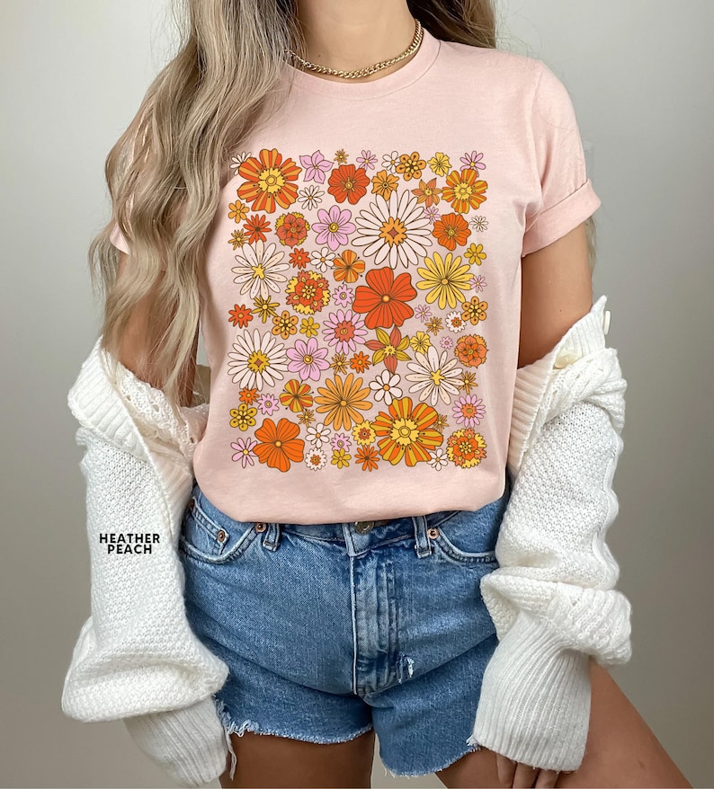May include: A pink t-shirt with a retro floral print featuring orange, yellow, and pink flowers. The shirt has a crew neck and short sleeves.