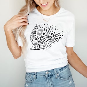 May include: A white t-shirt featuring a black and white graphic of a stylised bird with stars, moons, hearts, and arrows. The bird is flying with its wings outstretched.