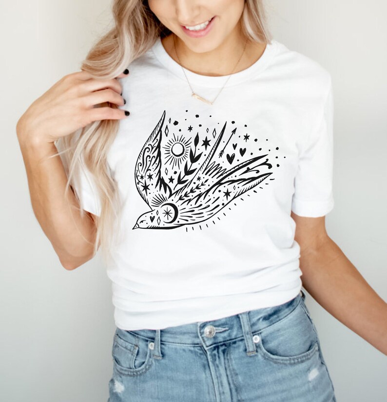 May include: A white t-shirt featuring a black and white graphic of a stylised bird with stars, moons, hearts, and arrows. The bird is flying with its wings outstretched.