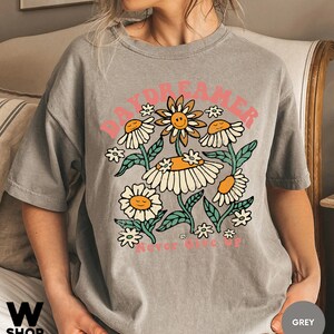 Puede incluir: Una camiseta gris con un gr&aacute;fico de un grupo de margaritas con caras sonrientes. El texto "Daydreamer" est&aacute; por encima de las margaritas y "Never Give Up" est&aacute; por debajo.