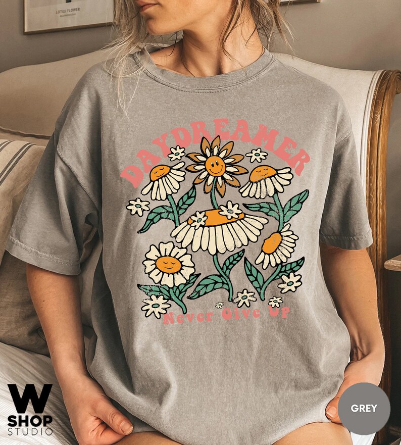 Puede incluir: Una camiseta gris con un gr&aacute;fico de un grupo de margaritas con caras sonrientes. El texto "Daydreamer" est&aacute; por encima de las margaritas y "Never Give Up" est&aacute; por debajo.