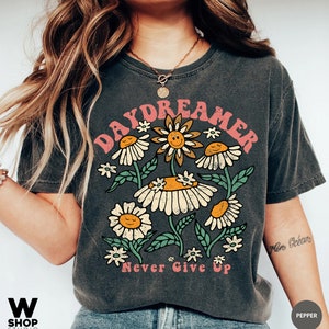Puede incluir: Una camiseta gris oscuro con un gr&aacute;fico vintage de flores y el texto "Daydreamer" y "Never Give Up".