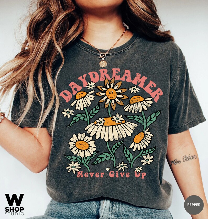 Puede incluir: Una camiseta gris oscuro con un gr&aacute;fico vintage de flores y el texto "Daydreamer" y "Never Give Up".