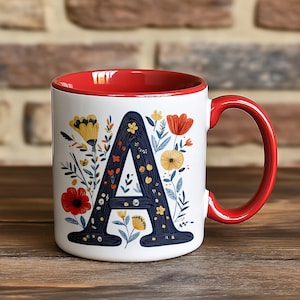 May include: White ceramic mug with a red interior and handle. The mug features a navy blue letter "A" filled with yellow and orange floral designs. The mug is decorated with additional floral elements.