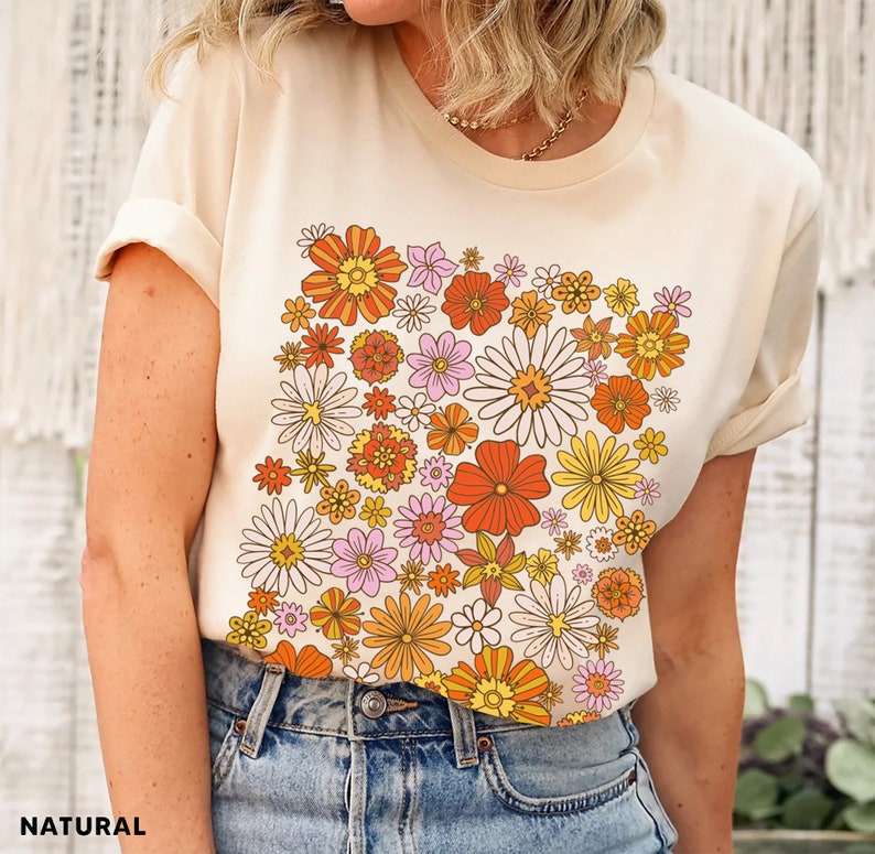 May include: A cream-colored t-shirt with a retro floral print featuring orange, yellow, pink, and white flowers. The flowers are arranged in a cluster on the front of the shirt.
