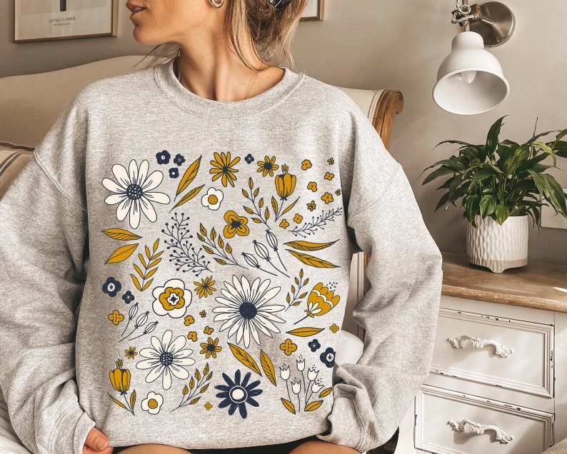 May include: A gray sweatshirt with a floral print featuring yellow, white, and blue flowers and leaves. The print is centered on the front of the sweatshirt.