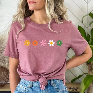 May include: A heather mauve t-shirt with a row of five colorful flowers printed on the front. The flowers are in shades of orange, yellow, pink, white, and green.