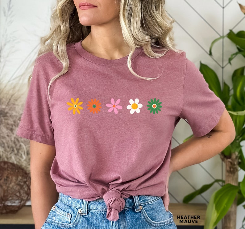 May include: A heather mauve t-shirt with a row of five colorful flowers printed on the front. The flowers are in shades of orange, yellow, pink, white, and green.