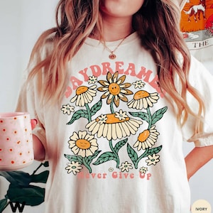 Puede incluir: Una camiseta blanca con un gr&aacute;fico rosa "Daydreamer" que presenta un grupo de margaritas amarillas con caras sonrientes. El texto "Never Give Up" est&aacute; impreso en rosa debajo de las margaritas.