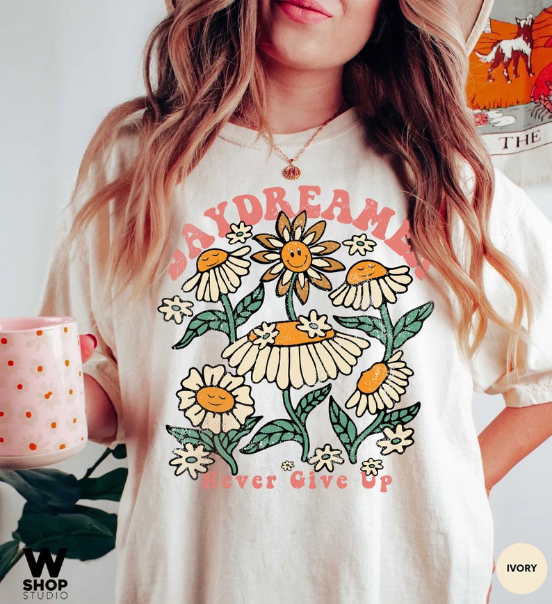 Puede incluir: Una camiseta blanca con un gr&aacute;fico rosa "Daydreamer" que presenta un grupo de margaritas amarillas con caras sonrientes. El texto "Never Give Up" est&aacute; impreso en rosa debajo de las margaritas.