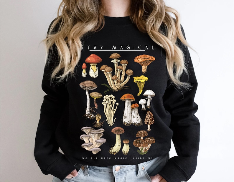 Op de afbeelding: Zwart sweatshirt met een paddenstoelprint en de tekst "Stay Magical" en "We all have magic inside us".
