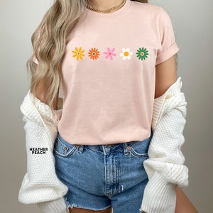 May include: A pink t-shirt with a row of five colorful flowers printed on the front. The flowers are yellow, orange, pink, white, and green.