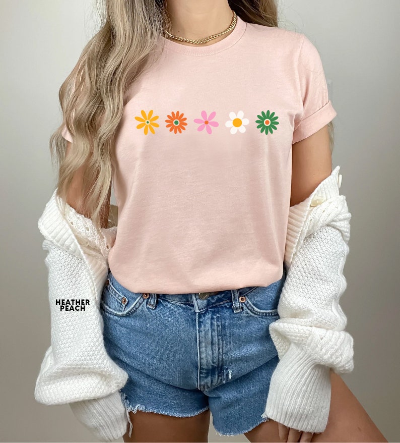 May include: A pink t-shirt with a row of five colorful flowers printed on the front. The flowers are yellow, orange, pink, white, and green.