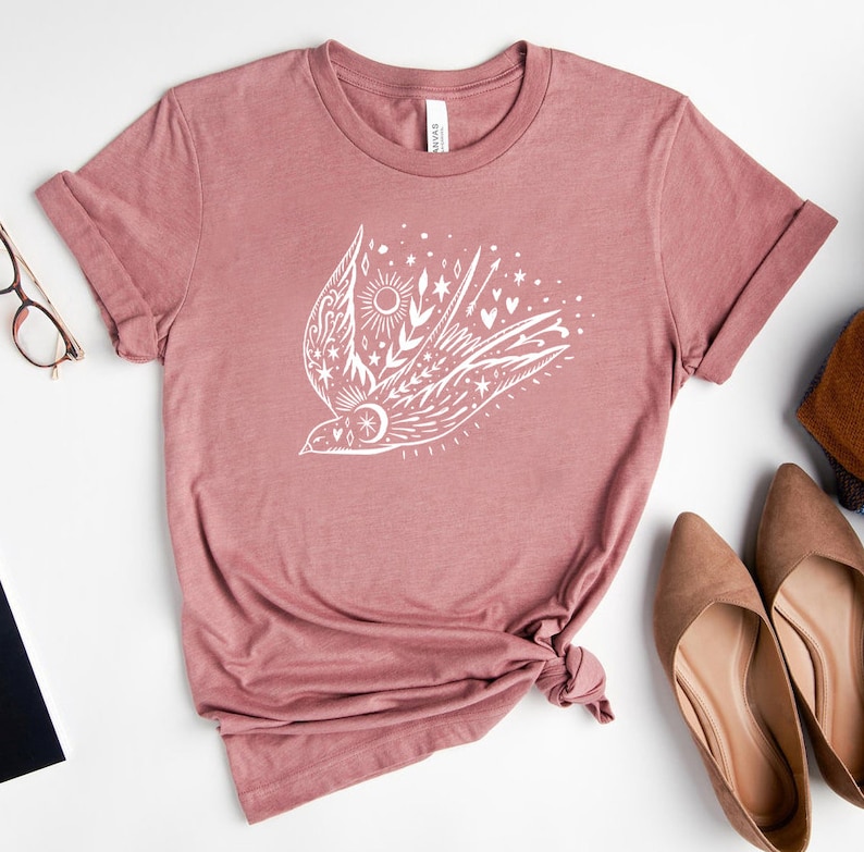 May include: A mauve t-shirt featuring a white graphic of a stylised bird with celestial elements, including stars, moons, and hearts.