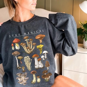 Op de afbeelding: Een donkergrijs sweatshirt met een paddenstoelprint en de tekst "STAY MAGICAL".