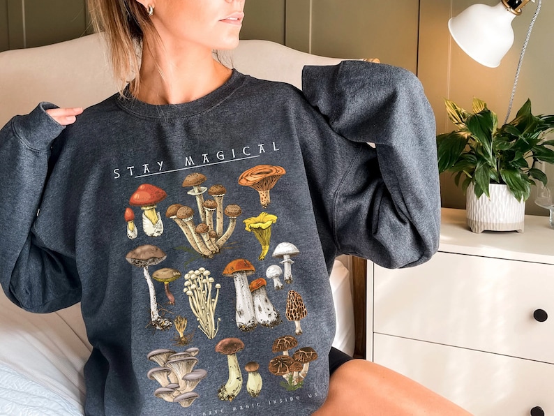 Op de afbeelding: Een donkergrijs sweatshirt met een paddenstoelprint en de tekst "STAY MAGICAL".