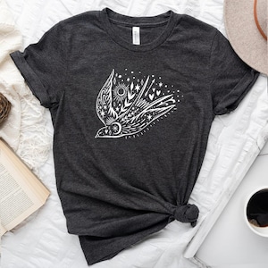 May include: A dark grey t-shirt featuring a white graphic of a stylised bird in flight, surrounded by stars, hearts, and a sun. The bird is adorned with floral details.