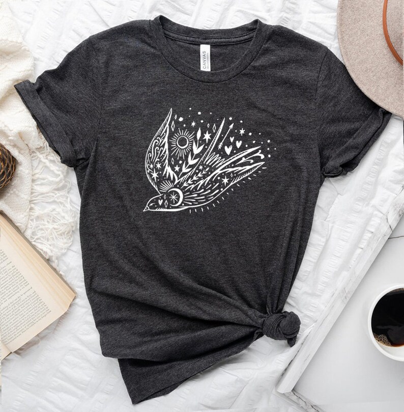 May include: A dark grey t-shirt featuring a white graphic of a stylised bird in flight, surrounded by stars, hearts, and a sun. The bird is adorned with floral details.