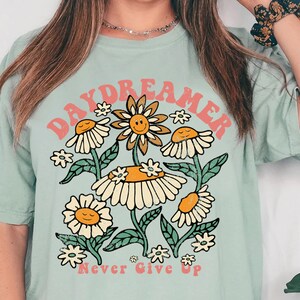 Puede incluir: Una camiseta azul claro con un dise&ntilde;o gr&aacute;fico retro que presenta el texto "Daydreamer" en rosa y naranja con un grupo de flores estilizadas y el texto "Never Give Up" en rosa.