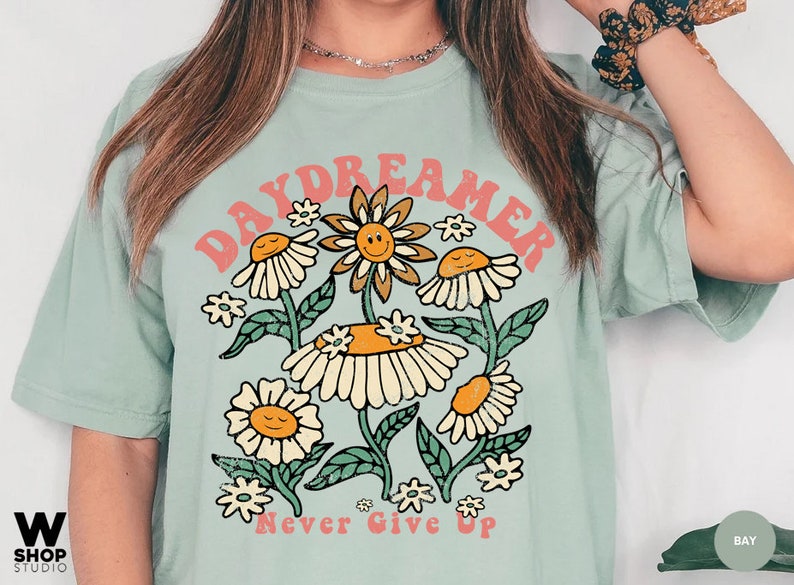 Puede incluir: Una camiseta azul claro con un dise&ntilde;o gr&aacute;fico retro que presenta el texto "Daydreamer" en rosa y naranja con un grupo de flores estilizadas y el texto "Never Give Up" en rosa.