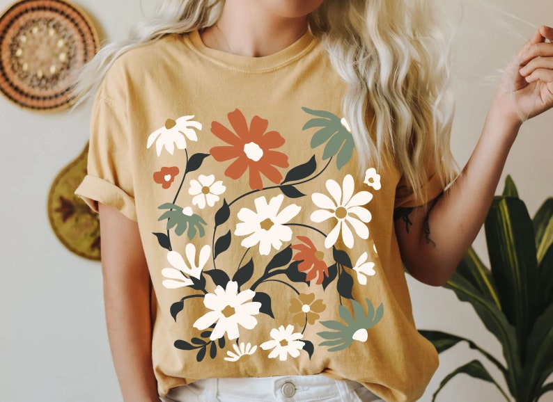 May include: Mustard yellow t-shirt featuring a floral design with white, rust, and sage green flowers. The graphic includes black stems and leaves. The shirt has short sleeves and a crew neck.