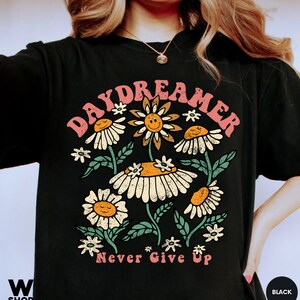 Puede incluir: Camiseta negra con un gr&aacute;fico de un arreglo de flores de margarita con caras sonrientes y el texto "Daydreamer" arriba y "Never Give Up" abajo.