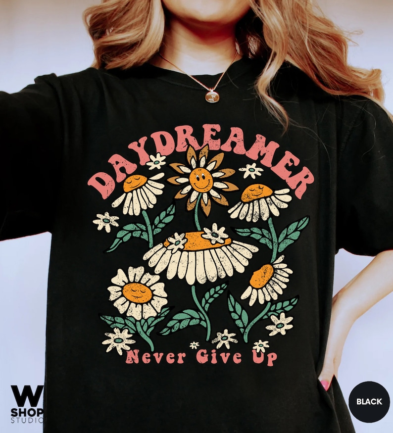 Puede incluir: Camiseta negra con un gr&aacute;fico de un arreglo de flores de margarita con caras sonrientes y el texto "Daydreamer" arriba y "Never Give Up" abajo.