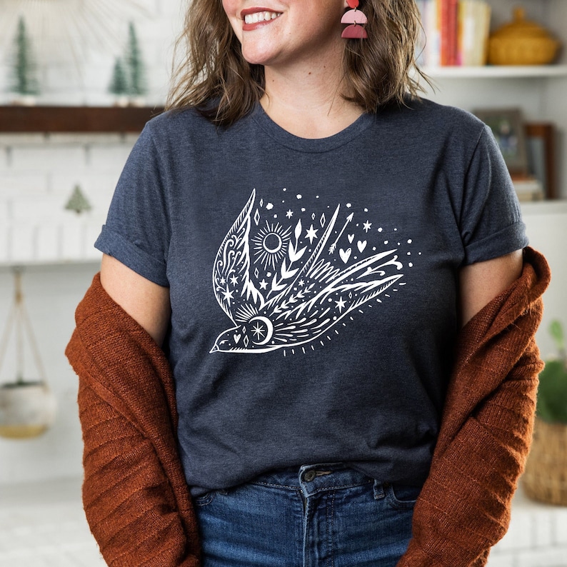 May include: A navy blue t-shirt featuring a white graphic of a stylised bird in flight, surrounded by stars, hearts and a sun. The bird is outlined in white.
