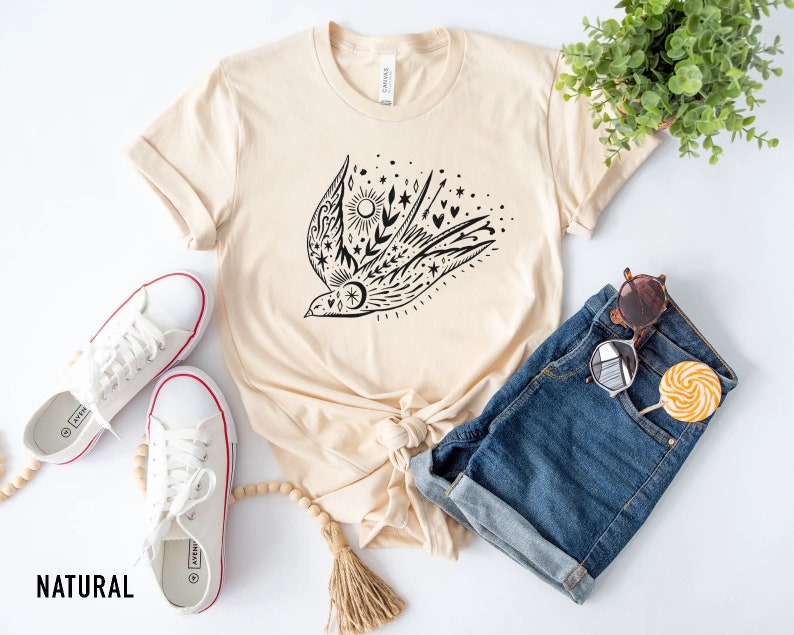 May include: A beige t-shirt featuring a black graphic of a bird with stars, hearts, and a sun. The shirt is laid out on a white surface with a pair of white trainers, a pair of blue denim shorts, sunglasses, and a lollipop.