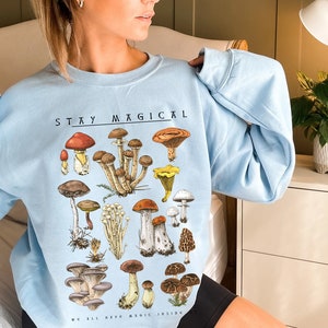 Op de afbeelding: Een lichtblauw sweatshirt met een paddenstoelprint en de tekst "Stay Magical We All Have Magic Inside".