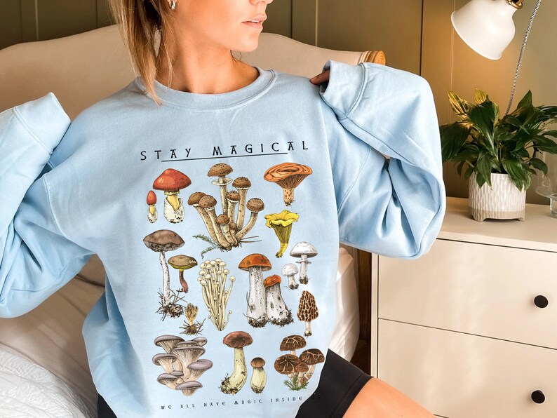 Op de afbeelding: Een lichtblauw sweatshirt met een paddenstoelprint en de tekst "Stay Magical We All Have Magic Inside".