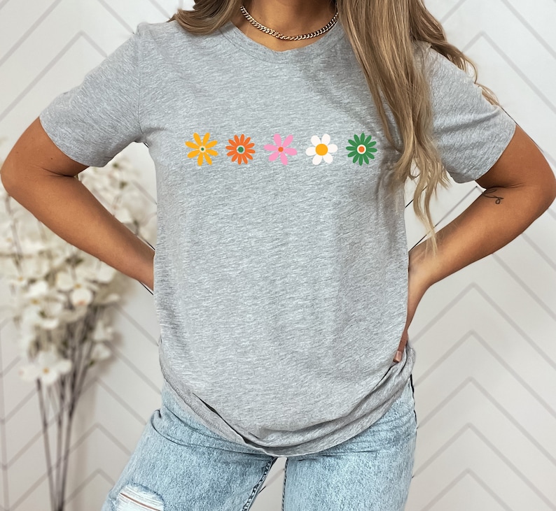 May include: A gray t-shirt with a row of five colorful flowers printed on the front. The flowers are orange, orange, pink, white, and green.