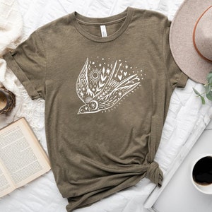 May include: A green t-shirt featuring a white graphic of a stylised bird in flight, surrounded by stars, hearts and a sun. The bird is surrounded by a floral design.