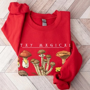 Op de afbeelding: Rood sweatshirt met een witte afbeelding van paddenstoelen en de tekst "STAY MAGICAL".